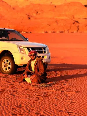 a man sitting in the desert next to a car at Wadi Rum Red Desert in Wadi Rum