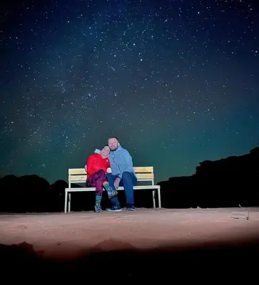 a man and woman sitting on a bench under the stars at Wadi Rum Mountains in Wadi Rum