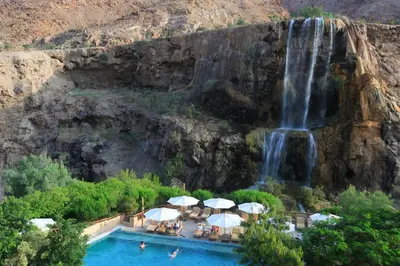 a waterfall and swimming pool with umbrellas in front of a waterfall at Ma'in Hot Springs in Sowayma